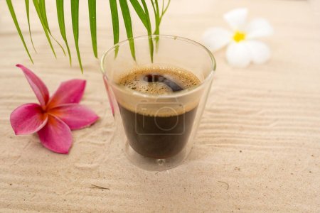 Photo for A transparent double glass mug with espresso coffee with white and pink Plumeria flowers on the sand - Royalty Free Image
