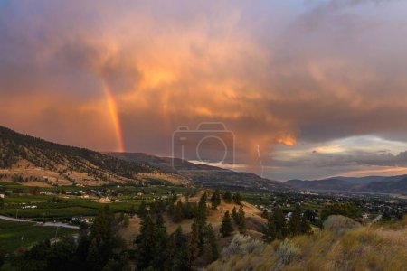Foto de Una vista de un arco iris en las nubes rojas y relámpagos blancos sobre las montañas y una ciudad. - Imagen libre de derechos