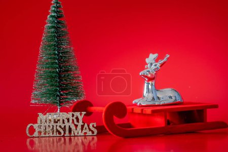 Foto de Un primer plano de un reno decorativo en miniatura en una matanza junto a un árbol de Navidad sobre un fondo rojo - Imagen libre de derechos