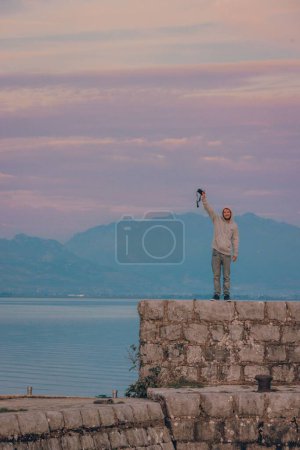 Téléchargez les photos : Un plan vertical d'un homme grimpé sur un mur de pierre et tenant une caméra avec un lac en arrière-plan entouré de collines - en image libre de droit