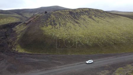 Photo for A drone shot of a white car going by a green moss-covered volcanic mountain and black lava field in Landmannalaugar, Iceland - Royalty Free Image