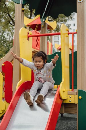 Téléchargez les photos : Plan vertical d'une petite fille glissant sur un toboggan rouge-jaune dans une aire de jeux - en image libre de droit
