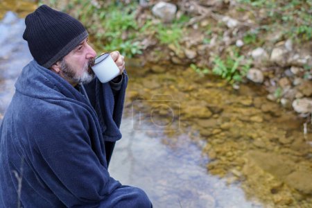 Téléchargez les photos : Homme barbu couvert d'une couverture buvant du café sur la rive de la rivière - en image libre de droit