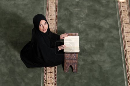 Photo for A high-angle shot of a Muslim woman in a black dress with hijab reading the Quran. - Royalty Free Image