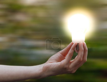 Foto de Un primer plano de la mano de una mujer sosteniendo una bombilla brillante sobre un fondo natural verde borroso - concepto de idea - Imagen libre de derechos