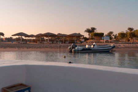 Téléchargez les photos : Paysage d'un bateau sur une baie au coucher du soleil à El-Gouna, Egypte avec parasols en paille dans la plage - en image libre de droit