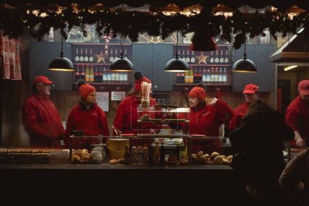 Foto de Un grupo de personas en uniformes rojos frente a una mesa en un puesto de Hot Dog en un mercado de Navidad - Imagen libre de derechos