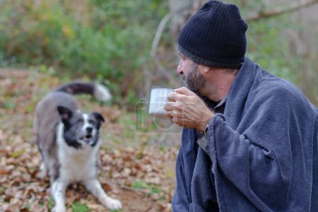 Foto de Barbudo cubierto con una manta bebiendo una taza de café con su perro en el fondo - Imagen libre de derechos