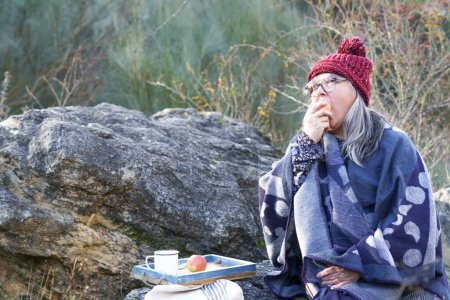 Foto de Mujer de pelo blanco con gorra roja comiendo una manzana sentada en una roca en el campo - Imagen libre de derechos