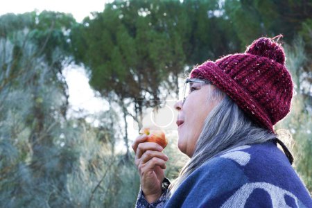Téléchargez les photos : Femme aux cheveux blancs avec chapeau rouge recouvert d'une couverture mangeant une pomme dans les montagnes - en image libre de droit