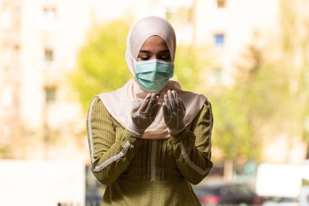 Téléchargez les photos : Une jeune femme musulmane priant à l'extérieur portant un masque protecteur et des gants - en image libre de droit