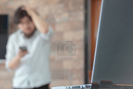 Photo for A laptop on a desk and a worried unrecognizable businessman out of focus in the background. Home Office. - Royalty Free Image