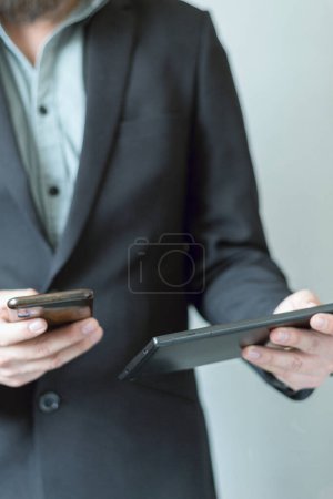 Foto de Un ejecutivo con un traje sin corbata sosteniendo un celular y una tableta. Un hombre de negocios que usa tecnología. Conexión multiplataforma. - Imagen libre de derechos