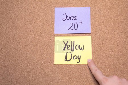 A hand points to a Yellow Day reminder sticky note placed on a cork board. June 20th