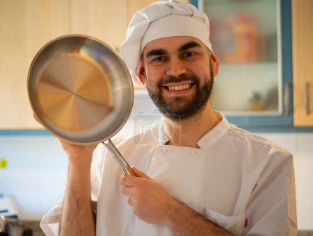 Téléchargez les photos : Portrait of a young bearded chef smiling while pointing a stainless pan looking at camera - en image libre de droit