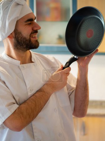 Téléchargez les photos : Portrait d'un jeune chef barbu souriant tout en montrant une casserole en acier inoxydable - en image libre de droit
