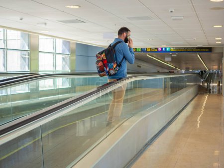 A young man on a moving walkway inside an airport using his phone