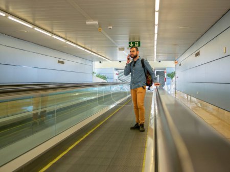 A young man on a moving walkway inside an airport using his phone