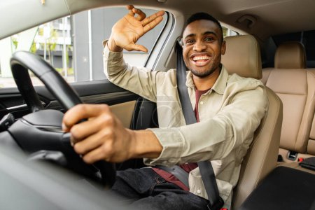 Junger afrikanisch-amerikanischer Mann lächelt beim Autofahren und winkt freundlich. Hochwertiges Foto