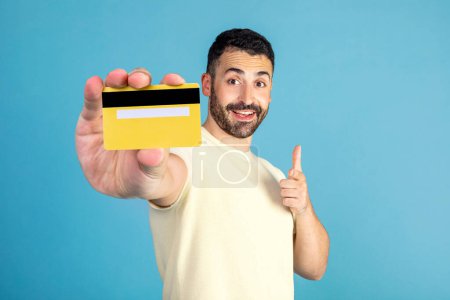 Foto de Positive man showing credit card, pointing at it and smiling, posing on blue background in studio. Money and finances concept - Imagen libre de derechos