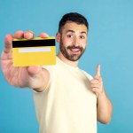 Positive man showing credit card, pointing at it and smiling, posing on blue background in studio. Money and finances concept