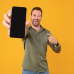 Excited caucasian man holding blank cellphone showing screen to camera pointing at it, recommending website or application, mockup banner