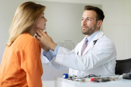 Photo for Professional male doctor examining thyroid gland of female patient during checkup in private clinic, sitting at desk in hospital office - Royalty Free Image