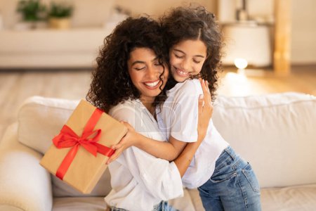 Photo for Mothers day concept. Adorable latin girl giving gift and embracing mom, mommy and daughter bonding together at home, sitting on sofa - Royalty Free Image