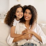 Happy latin girl hugging her beautiful mother and smiling together at camera, sitting on sofa in living room, adorable family embracing at home