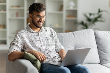 Photo for Positive hindu man freelancer working using laptop computer sitting on couch at home interior, guy surfing Internet, using modern technologies, free space - Royalty Free Image