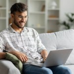 Positive hindu man freelancer working using laptop computer sitting on couch at home interior, guy surfing Internet, using modern technologies, free space