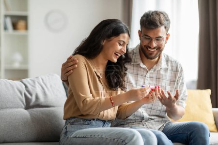 Happy indian spouses holding positive pregnancy test, sitting on couch at home together, couple celebrating awaiting baby, copy space