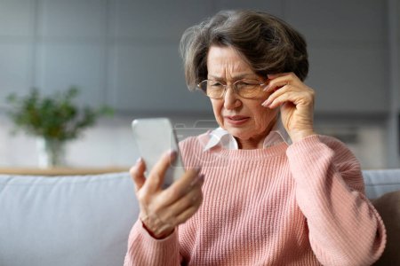 Téléchargez les photos : Femme âgée confus dans les lunettes qui louchent les yeux en lisant le message sur le téléphone portable, ayant des problèmes ophtalmiques avec la vision assis sur le canapé - en image libre de droit