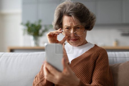 Femme européenne senior qui plisse les yeux en regardant l'écran du téléphone, portant des lunettes assis sur le canapé à la maison. Mauvaise vision problèmes de santé