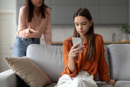 Photo for Angry mother shouting and arguing with teenage daughter over use of mobile phone, girl sitting on sofa at home, ignoring mom - Royalty Free Image