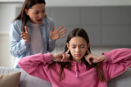 Photo for Strict mother scolding teenage daughter, mom lecturing teen girl, child covering ears with fingers, sitting on sofa. Parental harsh discipline, adolescent problem behavior - Royalty Free Image