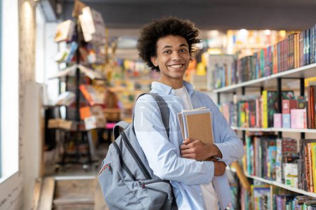 Happy black student guy with backpack immersed in studies, holding copybooks while standing in quiet library, showcasing dedication and focus on education