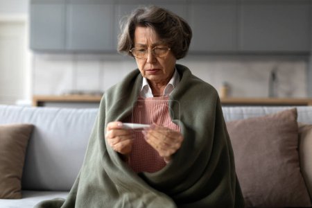 Téléchargez les photos : Malade femme âgée enveloppée dans une couverture regardant attentivement le thermomètre, se sentant malade et malade, assise sur le canapé dans le salon - en image libre de droit