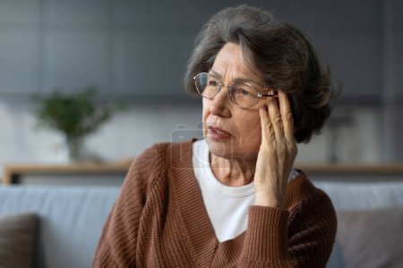Téléchargez les photos : Femme âgée bouleversée dans des lunettes assis sur le canapé à la maison, touchant ses temples et regardant de côté avec une expression troublée - en image libre de droit