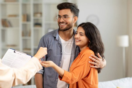 Real estate agent giving keys to young indian buyers, happy woman taking key of future new apartment, spouses buying new house