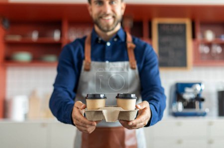 Téléchargez les photos : Barista mâle souriant dans un tablier tenant deux tasses jetables de café, travaillant dans un café. Concept de café à emporter - en image libre de droit