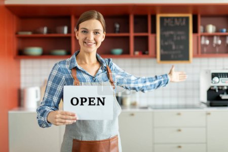 Retrato de la propietaria de la cafetería femenina gesto invitando a visitar y sosteniendo el cartel abierto, la señora sonriente de pie en el interior, espacio libre