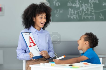 Alegre terapeuta del habla negra y niño pequeño aprendiendo y pronunciando letras durante la lección en la oficina