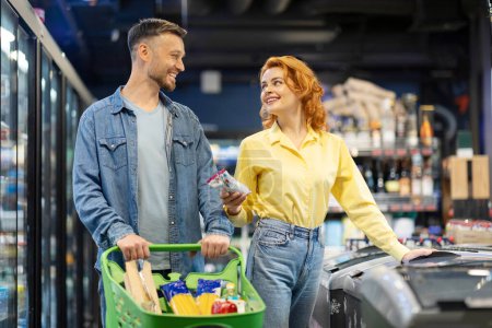 Téléchargez les photos : Heureux jeune couple faisant du shopping ensemble au supermarché, mari et femme debout au congélateur avec un chariot plein de nourriture - en image libre de droit
