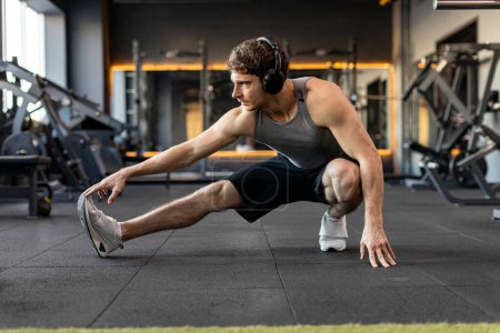 Muscular man in sportswear stretching on floor in empty gym, warming up before workout. Concept of bodybuilding, healthy lifestyle