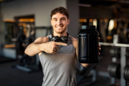 Photo for Sporty man demonstrating blank container for whey protein powder and pointing at it, recommending fitness supplements, mockup - Royalty Free Image