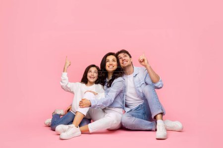 Familia feliz de tres mostrando descuentos de ventas de anuncios recomiendan la decisión de elección sentado aislado sobre fondo de color rosa