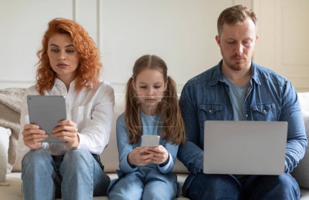 Gadget-Sucht. Eltern und Tochter bedienen sich verschiedener Gadgets, ignorieren einander, surfen mit gelangweiltem Gesichtsausdruck auf dem heimischen Sofa