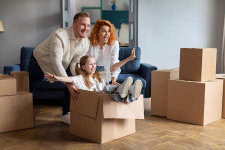 Photo for Happy family of three having fun and celebrating moving day, father and mother riding smiling daughter in cardboard box, fooling around in new flat - Royalty Free Image