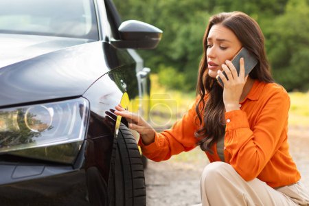 Traurige junge Frau sitzt neben zerkratztem Auto und ruft Ehemann um Hilfe, der Probleme mit neuem Fahrzeug hat